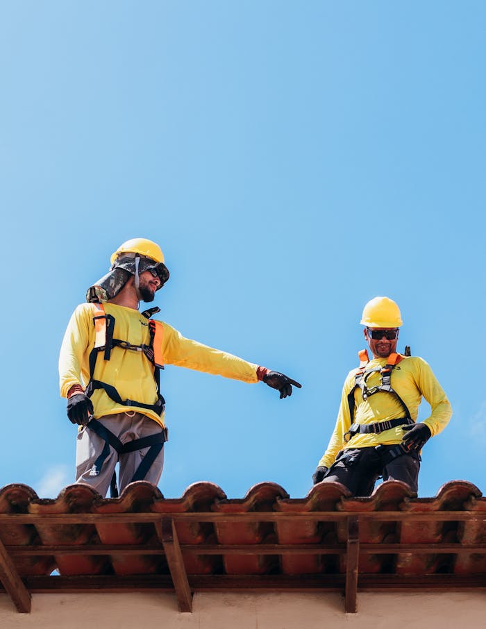 construction-workers-collaborating-on-rooftop-28812511 Two construction workers in safety gear discussing roofing project under clear blue sky.